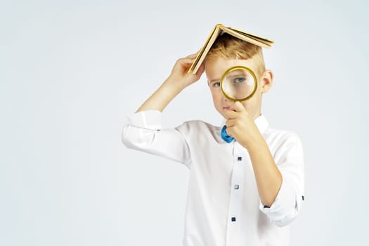 The schoolboy put the book on his head and looks through a magnifying glass. Isolated background. Education concept