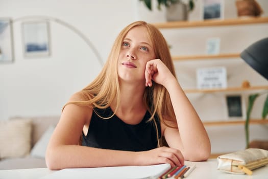 Sitting by the table. Female teenager with blonde hair is at home at daytime.