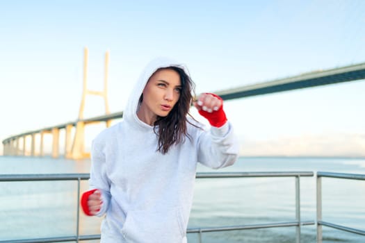 Woman boxing with her hands wrapped in red boxing tapes bridge and blue sky background. Middle age female athlete fighter training outside dressed hoodie.