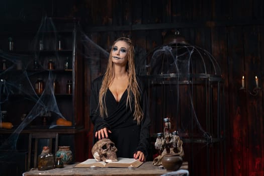 Halloween concept. Witch portrait close up with dreadlocks holding human scull in hand dressed black hood standing dark room occultism religion Caucasian female necromancer