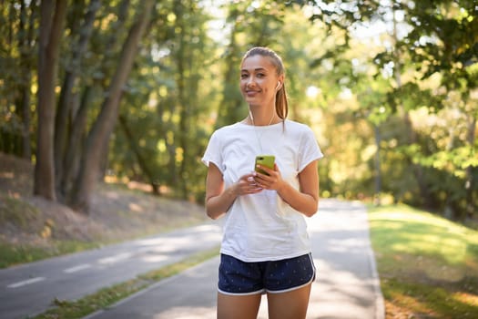 Woman putting on earphones to listen music before jogging summer park Caucasian female preparing to run sunny morning Runner ready training holdin smartphone hand Healthy lifestyle positive concept