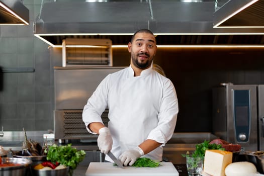 Handsome young African chef standing in professional kitchen in restaurant preparing a meal of meat and cheese vegetables. Portrait of man in cook uniform Cuts dill with a metal knife.