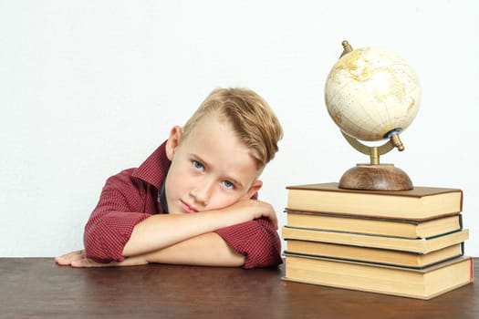 Education concept. A tired schoolboy sits at the table, put his head on his hands. On the table there are books, a globe and an alarm clock.