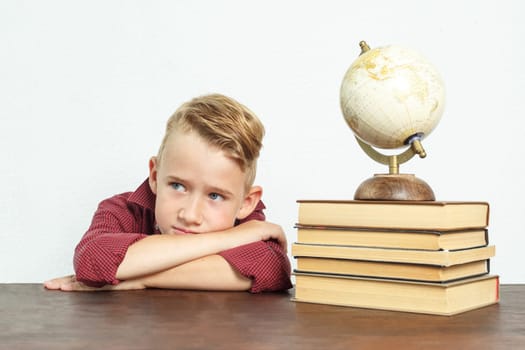 Education concept. A tired schoolboy sits at the table, put his head on his hands. On the table there are books, a globe and an alarm clock.