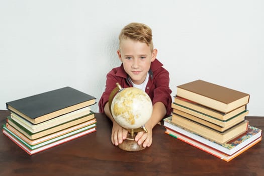 Education concept. The schoolboy sits at the table, shows the globe. On the table there are books, a globe and an alarm clock.