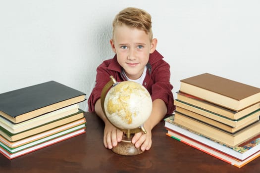 Education concept. The schoolboy sits at the table, shows the globe. On the table there are books, a globe and an alarm clock.