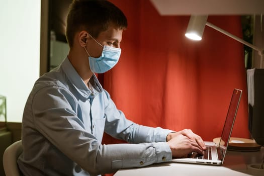 a young Caucasian man in a light shirt sits at a table in the evening at home wearing a protective medical mask and a mask and works at a laptop, remote work at home online