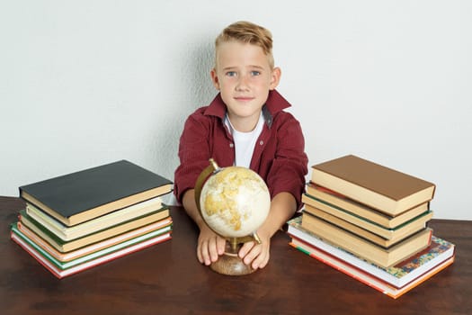 Education concept. The schoolboy sits at the table, shows the globe. On the table there are books, a globe and an alarm clock.