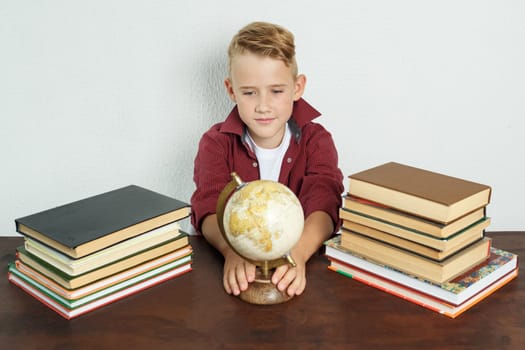 Education concept. The student sits at the table and looks at the globe holding on to it with his hands. On the table there are books, a globe and an alarm clock.