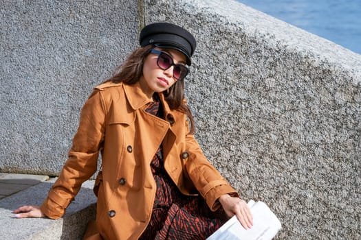 Young woman in sunglasses of Caucasian ethnicity sits on the steps on the embankment in a black cap and brown jacket with a newspaper in her hand