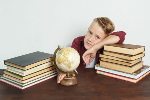 Education concept. The schoolboy sits at the table, shows the globe with his elbows on the books. On the table there are books, a globe and an alarm clock.