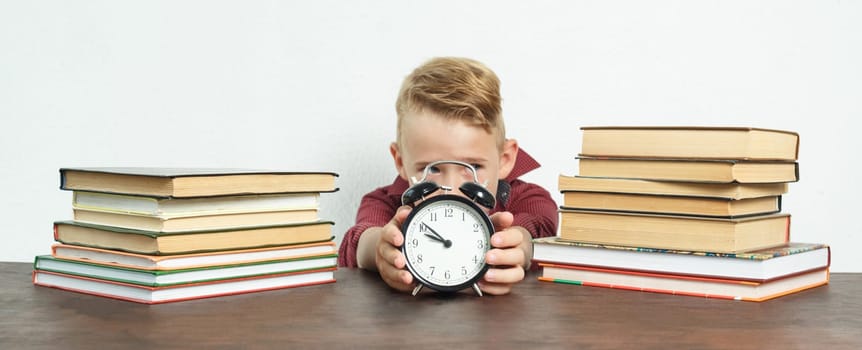 Education concept. The schoolboy sits at the table and holds an alarm clock in his hands. Sharpness of the image on the alarm clock