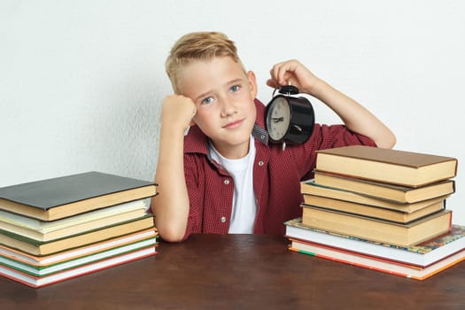 Education concept. The schoolboy sits at the table and holds an alarm clock in his hands.