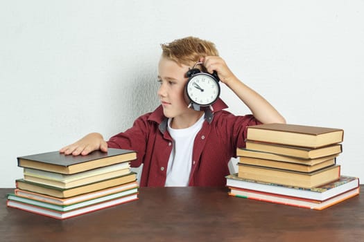 Education concept. The schoolboy sits at the table and holds an alarm clock in his hands.