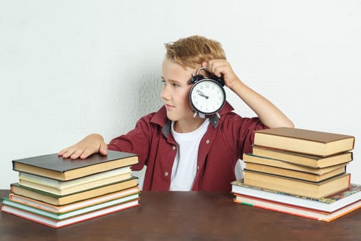 Education concept. The schoolboy sits at the table and holds an alarm clock in his hands.