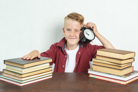 Education concept. The schoolboy sits at the table and holds an alarm clock in his hands.