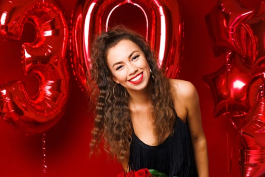 Portrait of happy 30 year old woman on red background with red balloons. A beautiful girl of Caucasian appearance is celebrating her anniversary. Balloons in the form of numbers 30 and stars.