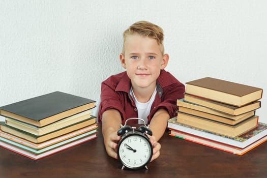 Education concept. The schoolboy sits at the table and holds an alarm clock in his hands.