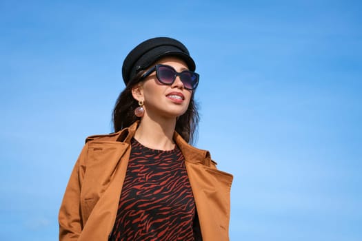 Female portrait of a beautiful young woman of Caucasian appearance in a black cap against a blue sky in sunglasses and a jacket