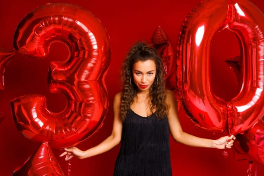 Valentine Beauty girl with red balloon. Beautiful Happy Caucasian Young woman Holiday party, birthday. Joyful model - Image. On a red background, red balloons in the form of numbers 30