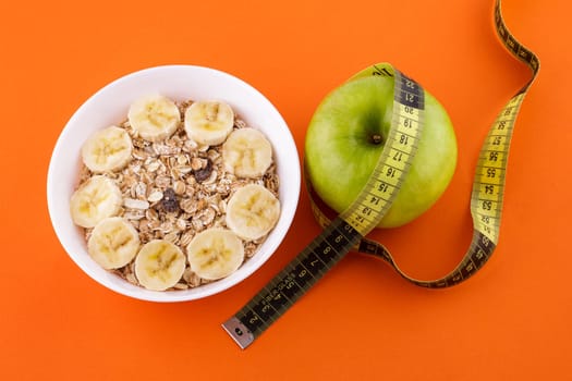 muesli with banana in a white plate on an orange background, green apple with a yellow measuring tape. Healthy food and diet concept