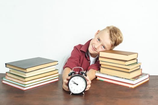 Education concept. The schoolboy sits at the table and holds an alarm clock in his hands.