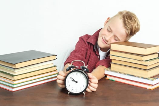 Education concept. Pupil boy sits at a table with books and holds an alarm clock in his hands.