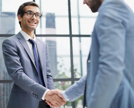 Businessmen shake hands, standing near a large office window