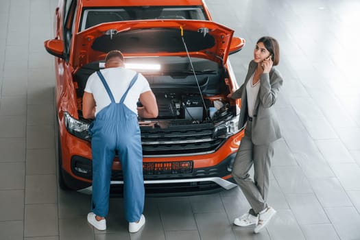 Woman in stylish clothes. Man repairing woman's automobile indoors. Professional service.