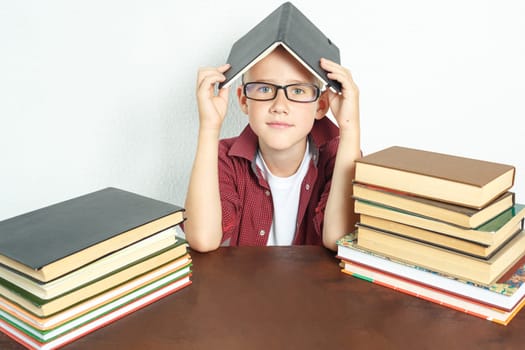 Education concept. A pupil boy sits at a table with books, holding an open book on his head.