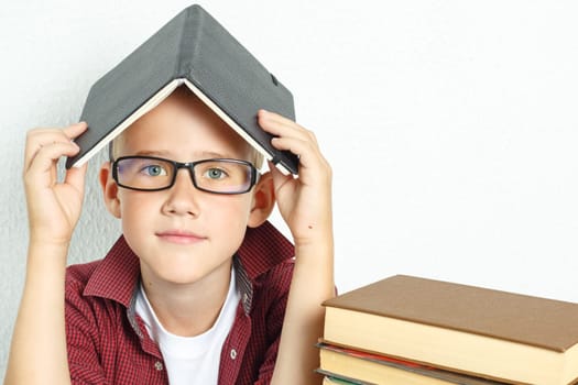Education concept. A pupil boy sits at a table with books, holding an open book on his head.