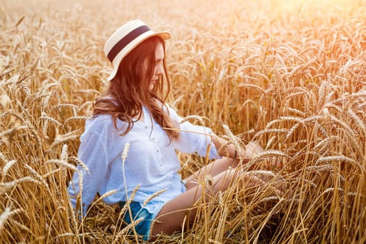 Happy girl sits in wheat field in straw hat. Cute young woman of Caucasian ethnicity in casual clothes enjoys ripe golden wheat in a field. Free woman concept. selective focus