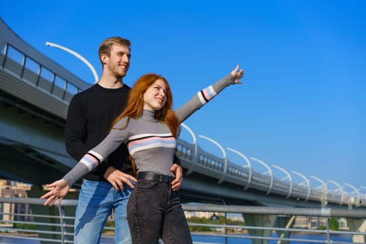 happy young couple man and woman with long red hair, stand against the background of a blue sky and a bridge in casual clothes and smile. Cheerful guy and Caucasian girl on a sunny day