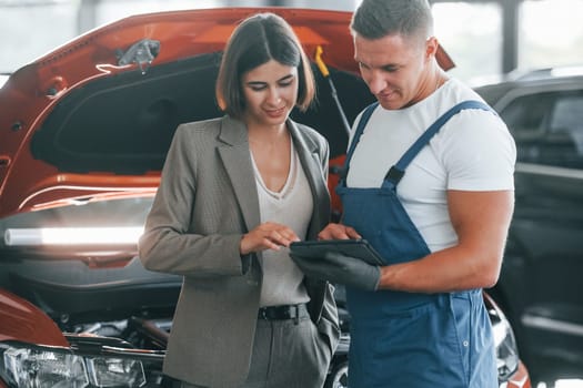 Woman in stylish clothes. Man repairing woman's automobile indoors. Professional service.