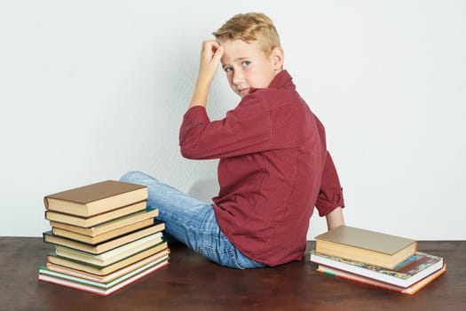 The schoolboy sits on the table near the books and holds his head with his hand, turning his back. Education concept