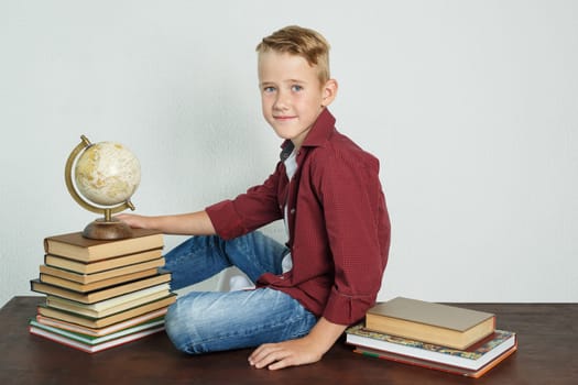 A schoolboy sits at a desk near books and holds a globe with his hand. Education concept