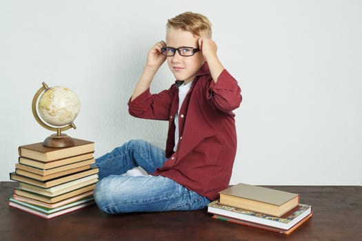 A schoolboy sits on a table near books and a globe, straightens his glasses. Education concept