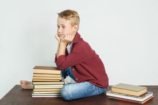 A schoolboy sits on the table near the books, resting his elbows on them and looking away. Education concept