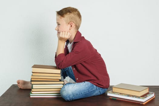 A schoolboy sits on the table near the books, resting his elbows on them and looking away. Education concept