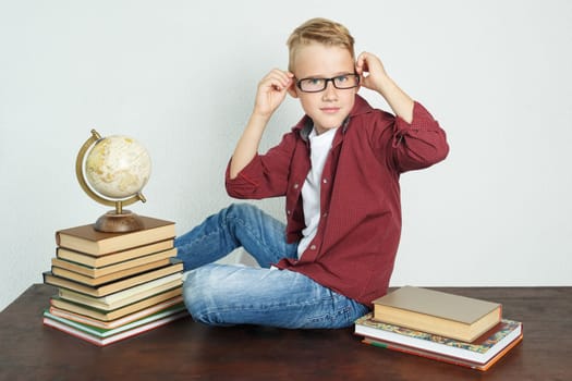 A schoolboy sits on a table near books and a globe, straightens his glasses. Education concept