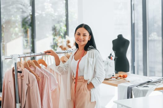 Standing and smiling. Seamstress is in her office that is with different clothes.