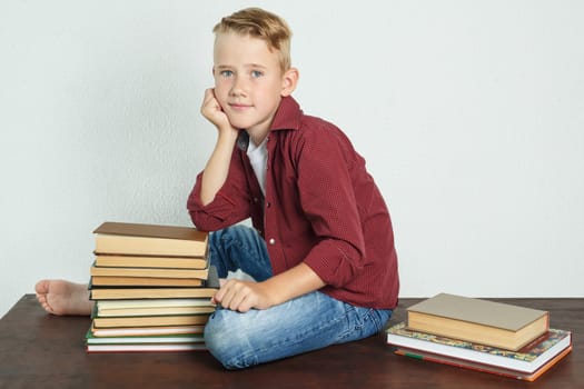 A schoolboy sits on the table near the books, leaning his elbows on them and looks into the cameras. Education concept