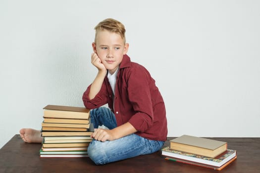 A schoolboy sits on the table near the books, resting his elbows on them and looking away. Education concept