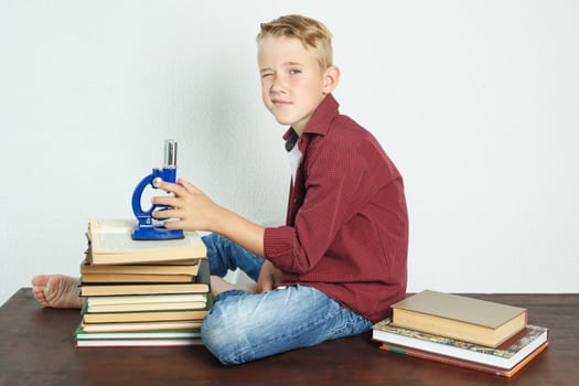 A schoolboy sits at a table near books, holds a microscope in his hands and looks at the camera. Education concept