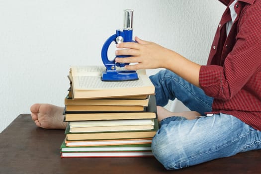 A schoolboy sits on a table near books and holds a microscope in his hands. Education concept
