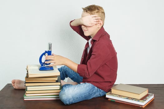 A schoolboy is sitting at a table near books, holding a microscope in his hands, covering his face with his hand. Education concept