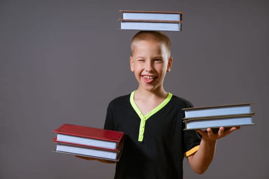 little blond boy in a black T-shirt hold stacks of books in your hands and on your head. the child is looking at the camera, isolated on a gray background.