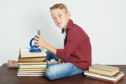 A schoolboy sits at a table near books, holds a microscope in his hands and looks at the camera. Education concept