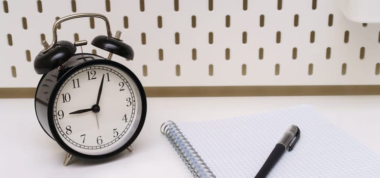 a black alarm clock, notepad, pen are placed on a white background of the table. five minutes past nine