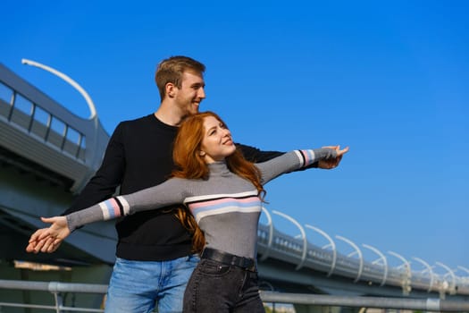 happy young couple man and woman with long red hair, stand against the background of a blue sky and a bridge in casual clothes and smile. Cheerful guy and Caucasian girl on a sunny day